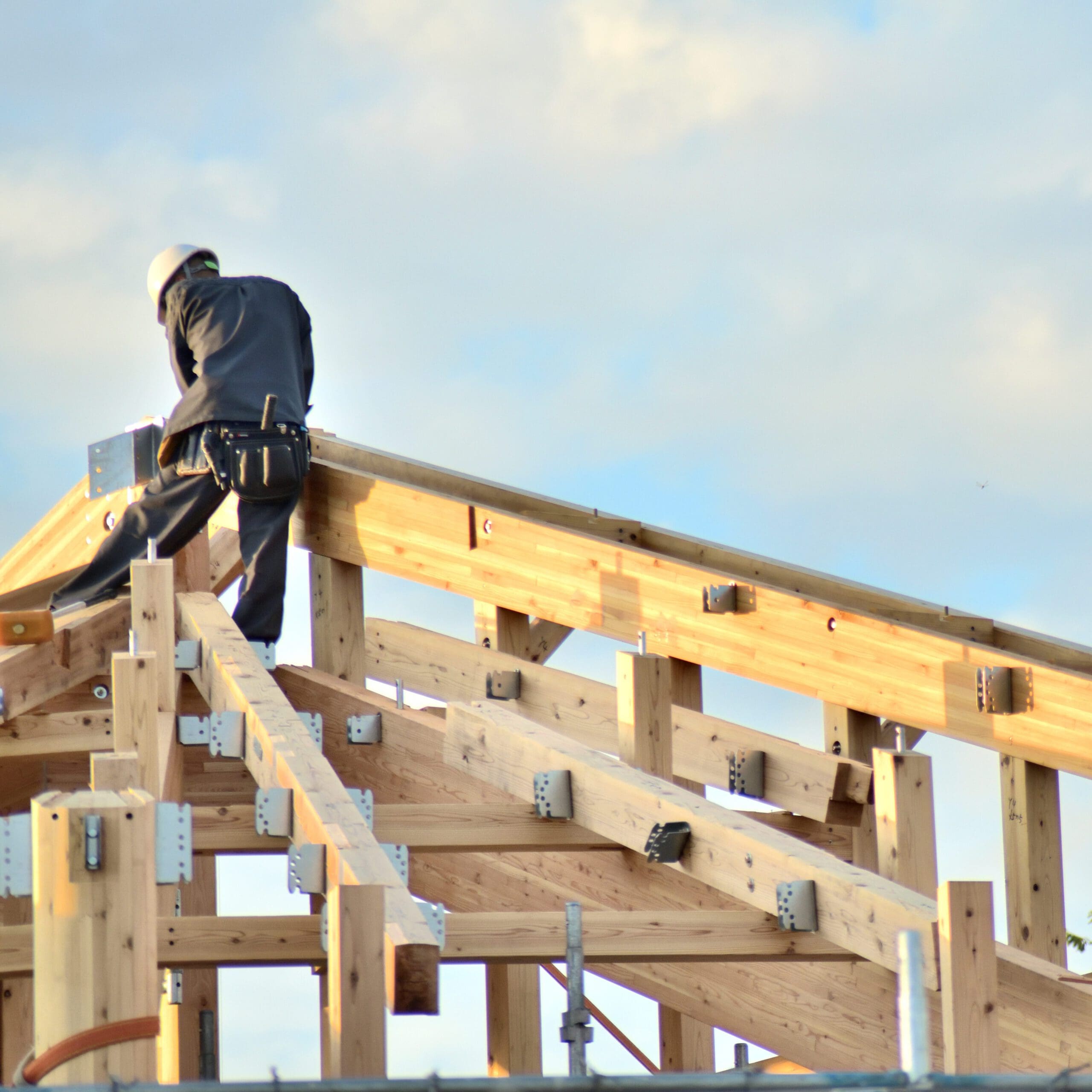 A construction worker wearing a hard hat and tool belt works on the wooden frame of a roof under a cloudy sky.