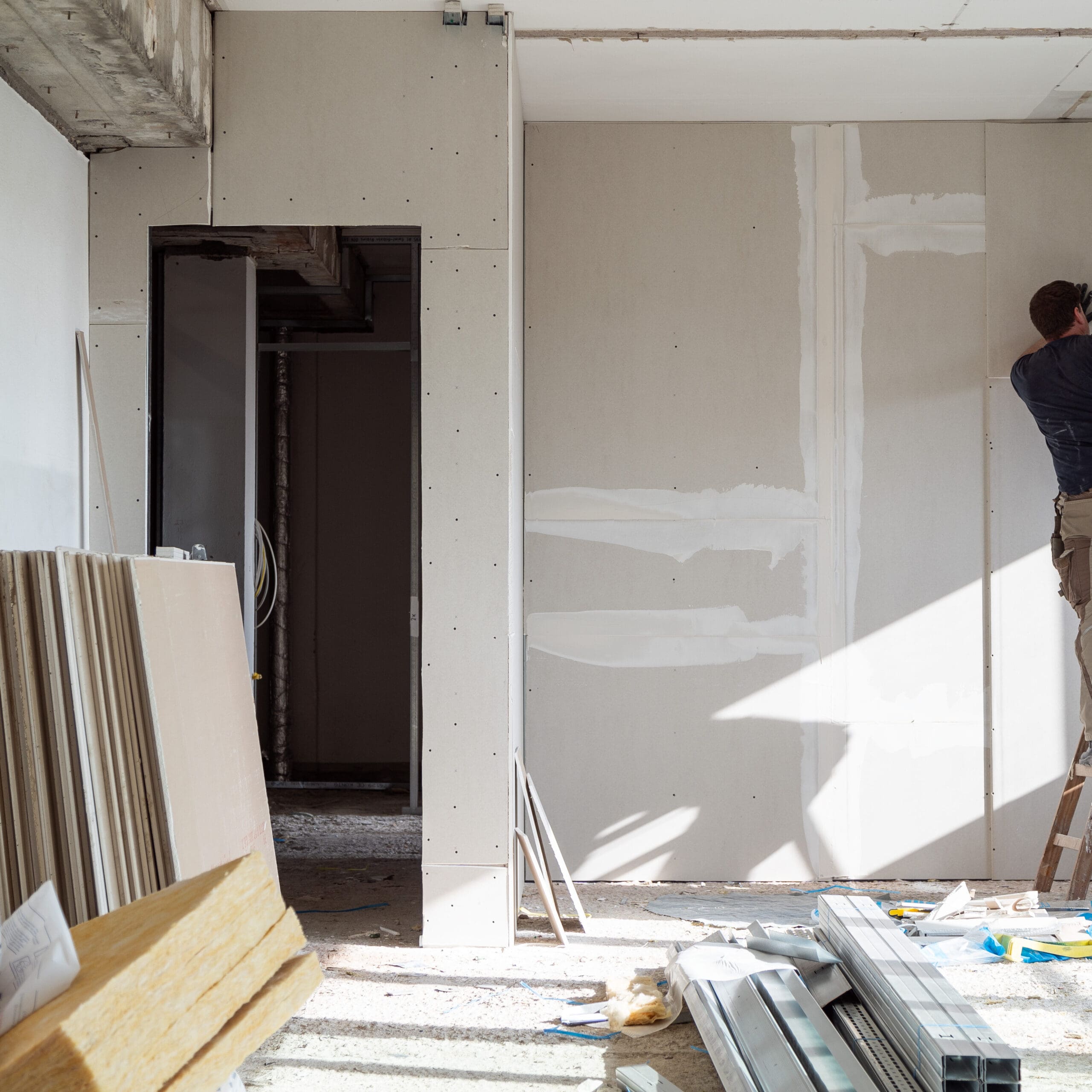 A construction worker stands on a ladder, installing drywall panels in a partially renovated room. Several stacks of drywall sheets and metal framing materials are scattered on the concrete floor. Sunlight streams in, illuminating the workspace.