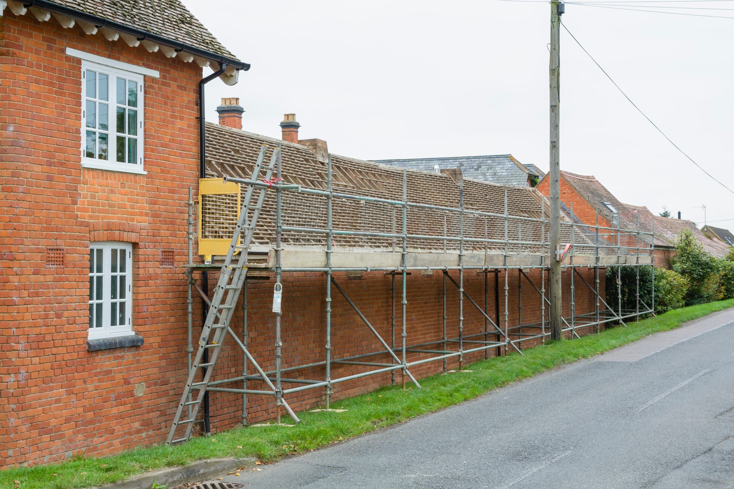 A long scaffolding structure is set up against a red brick building. A ladder leans against the scaffolding. The building, partially covered, is alongside a road with a telephone pole nearby. The sky is overcast.