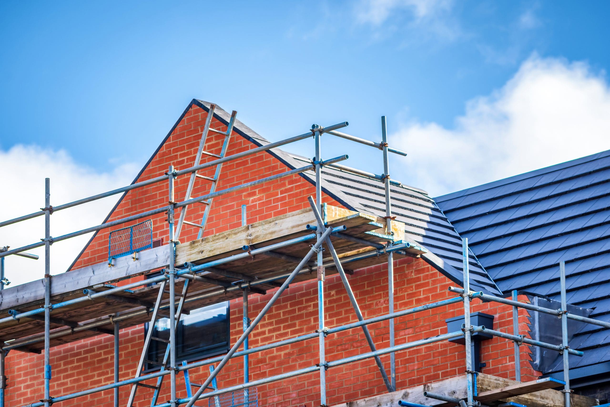 A brick house under construction with scaffolding on the roof. The roof is partially covered with dark blue tiles. The sky is bright blue with a few clouds.