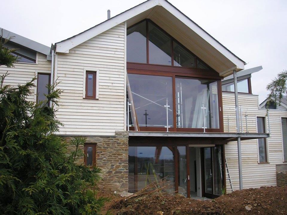 Modern two-story house with large glass windows and wooden siding. The building features a stone lower section, sloped roofs, and is surrounded by trees and soil. A ladder leans against the structure, indicating ongoing construction or maintenance.
