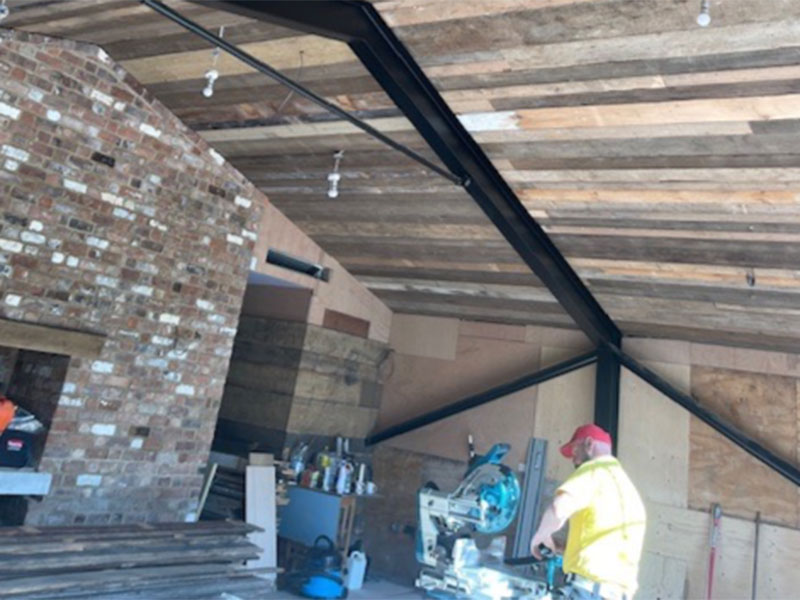 A person wearing a red cap and yellow vest operates a saw in a workshop with exposed wooden beams and brick walls. Various tools and materials are scattered around the space.