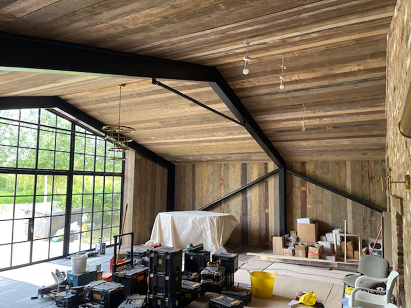 A spacious room under construction features a wooden ceiling with exposed metal beams, large windows, and various construction tools and materials scattered throughout. A covered object and boxes are on the floor, with an outdoor view visible.