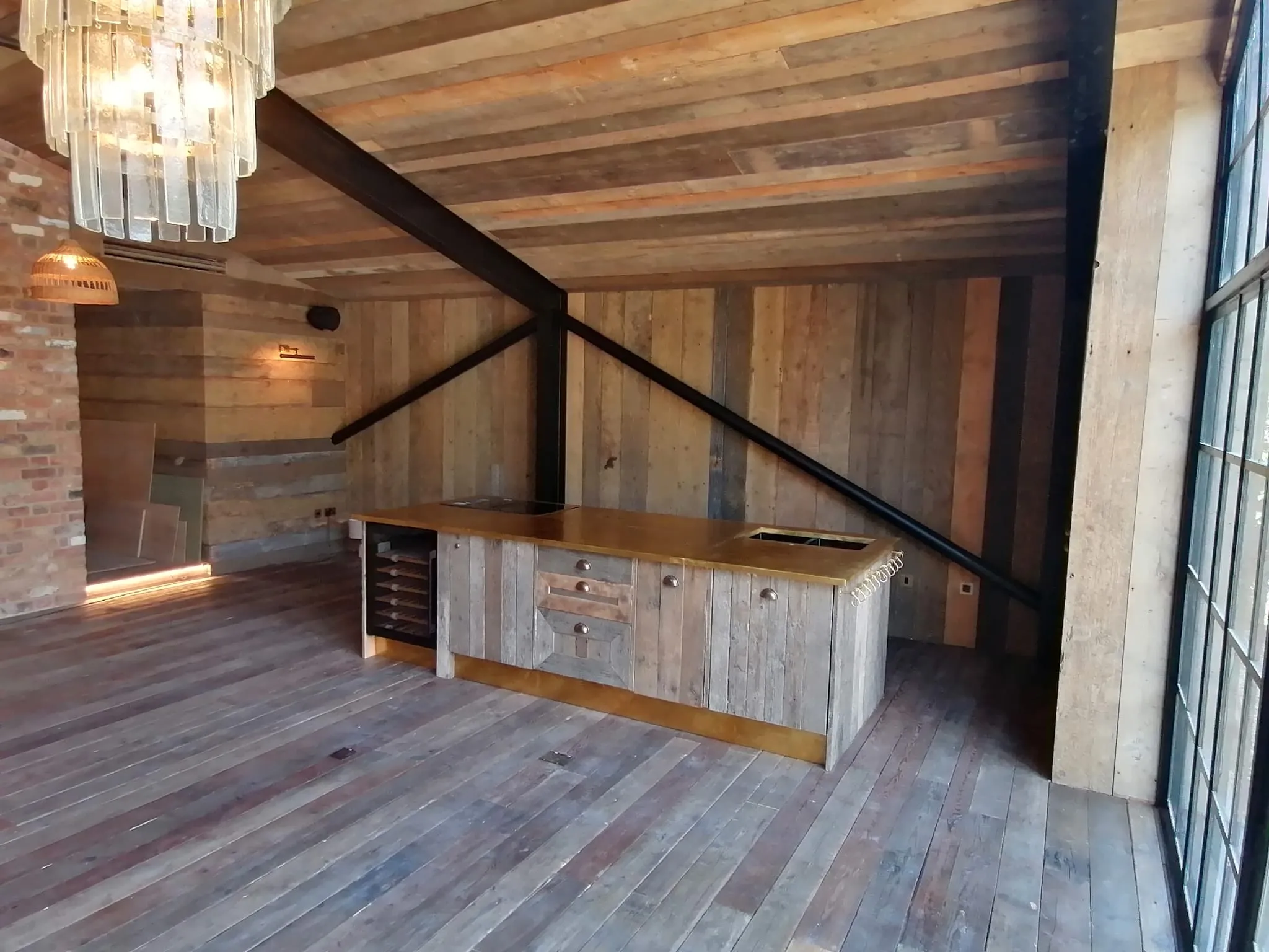 A rustic, modern kitchen with a central wooden island, large windows, and exposed beams. The room features wooden floors and walls, and a unique chandelier hanging from the ceiling, creating a cozy yet contemporary atmosphere.