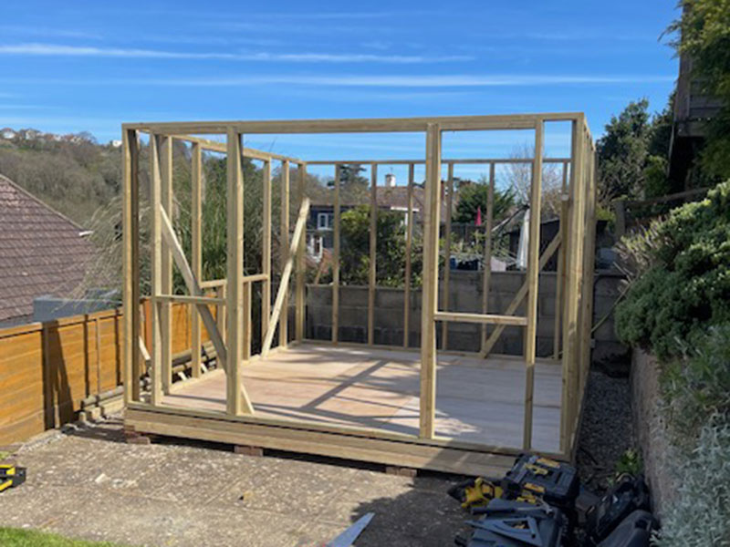 Wooden frame in the process of being constructed on a patio area with some tools on the ground nearby. The structure is set against a backdrop of houses and trees under a clear blue sky.
