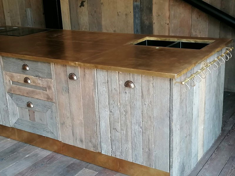 A rustic kitchen island with a wooden base and brass countertop. It features drawers, a small sink cutout, and a row of hooks on the side for hanging utensils. The wooden panels have a weathered, vintage look.