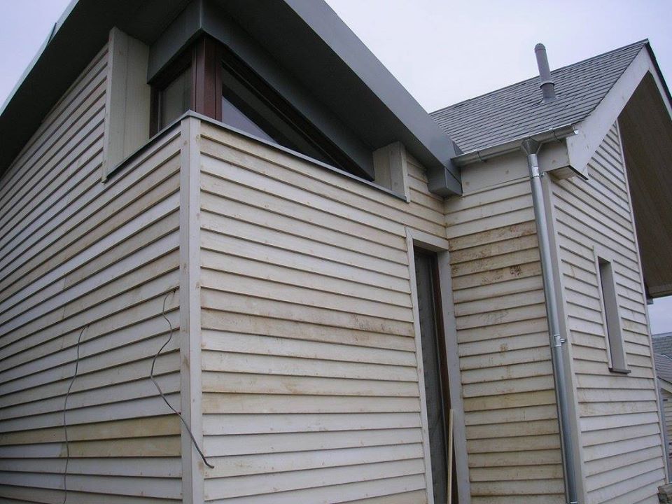 A close-up of a house's exterior corner featuring light-colored wooden siding with visible dirt streaks. The roof has gray shingles and a visible gutter system. A narrow vertical window is present near the corner.