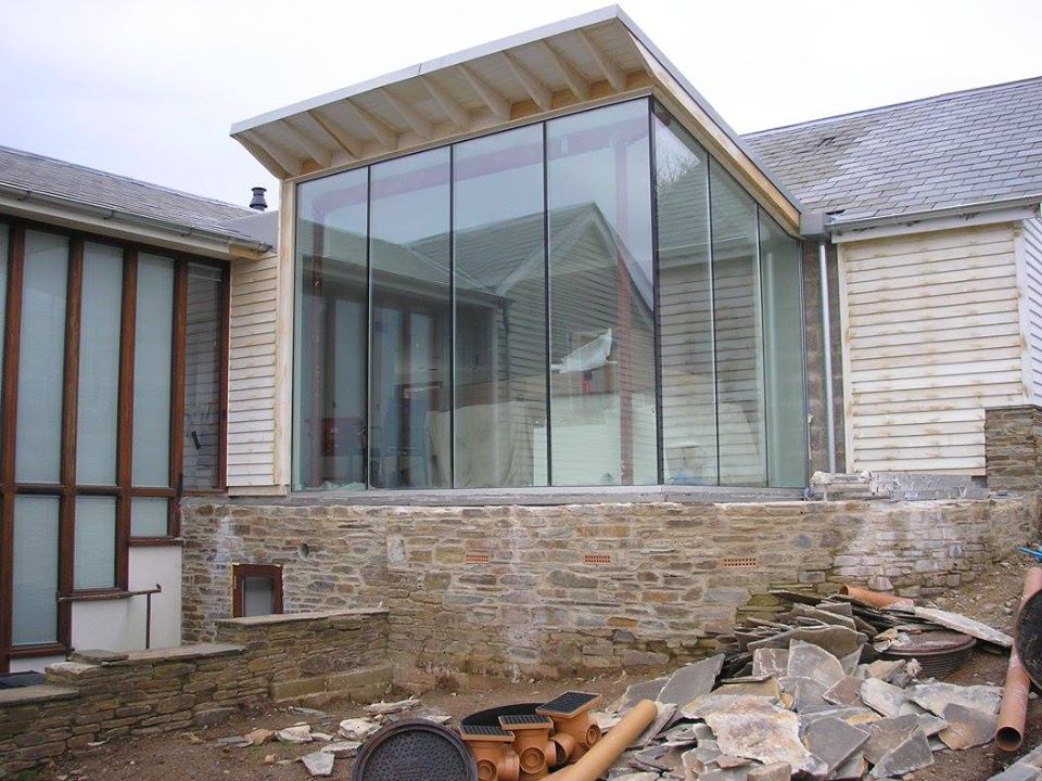A modern house extension under construction with large floor-to-ceiling glass windows. The building features a mix of stone and wood. Construction materials and debris are scattered in the foreground.