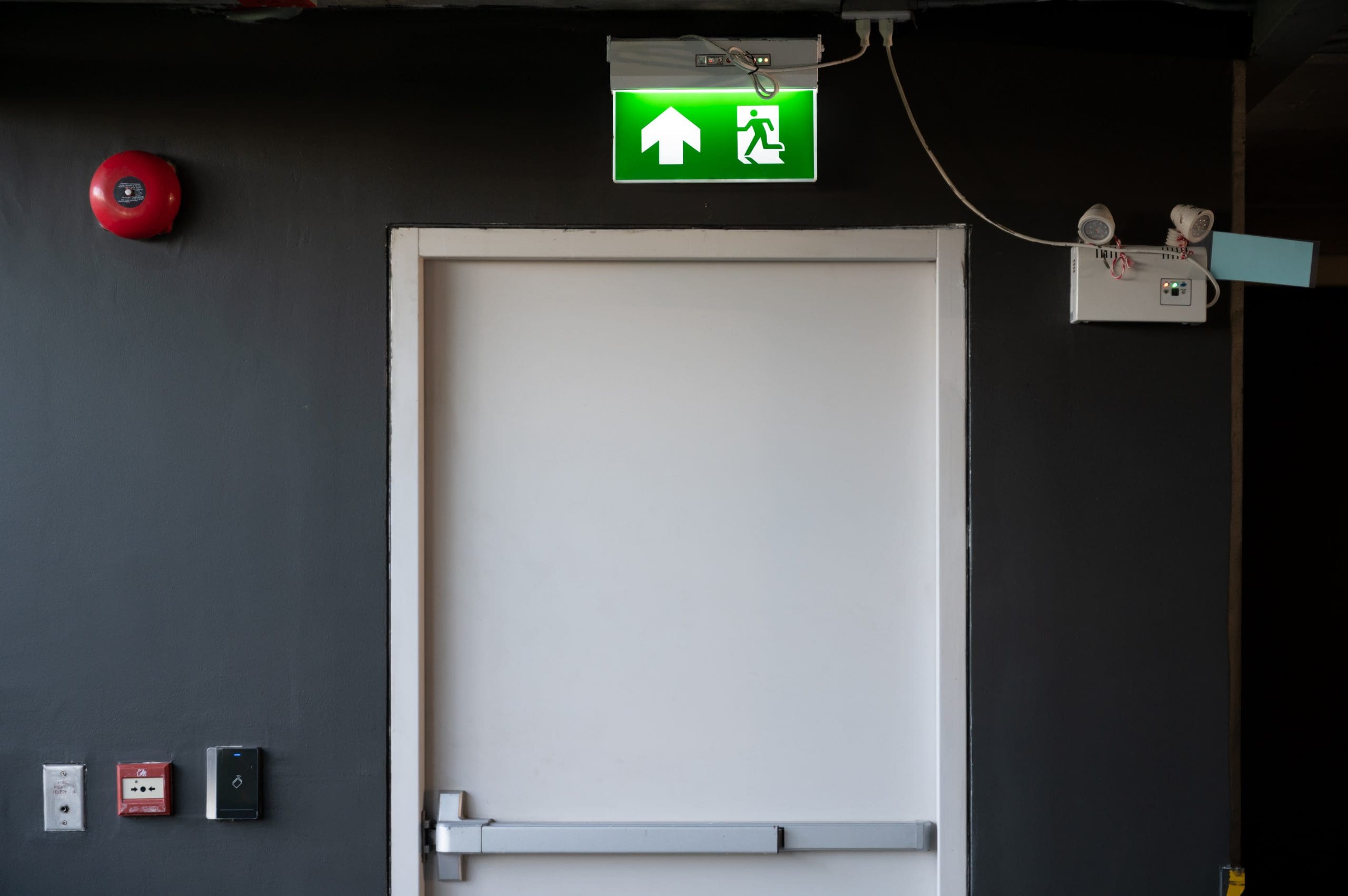 Closed white door in a dark hallway with a green illuminated emergency exit sign above, showing an arrow pointing up and a figure running. A red fire alarm and a security keypad are mounted on the left wall.