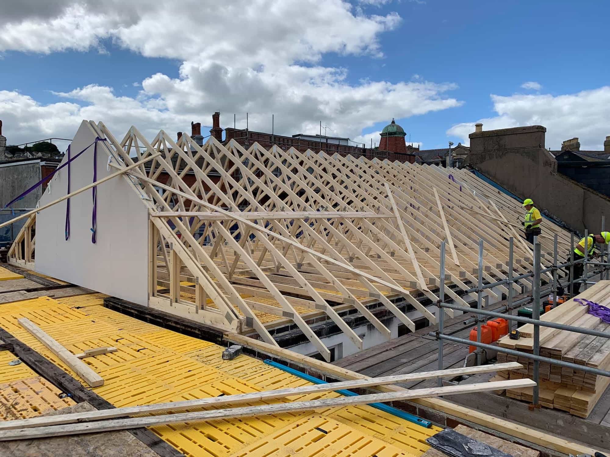 Construction workers in safety gear work on a building site, assembling a large wooden roof frame with exposed trusses. Scaffolding and materials surround the area under a partly cloudy sky.