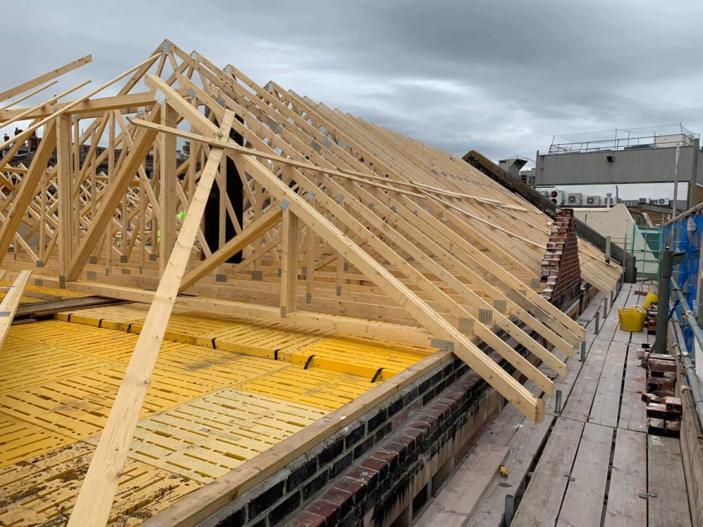 Wooden roof trusses and beams being installed on a building under construction, with scaffolding and tools visible on the right side under a cloudy sky.