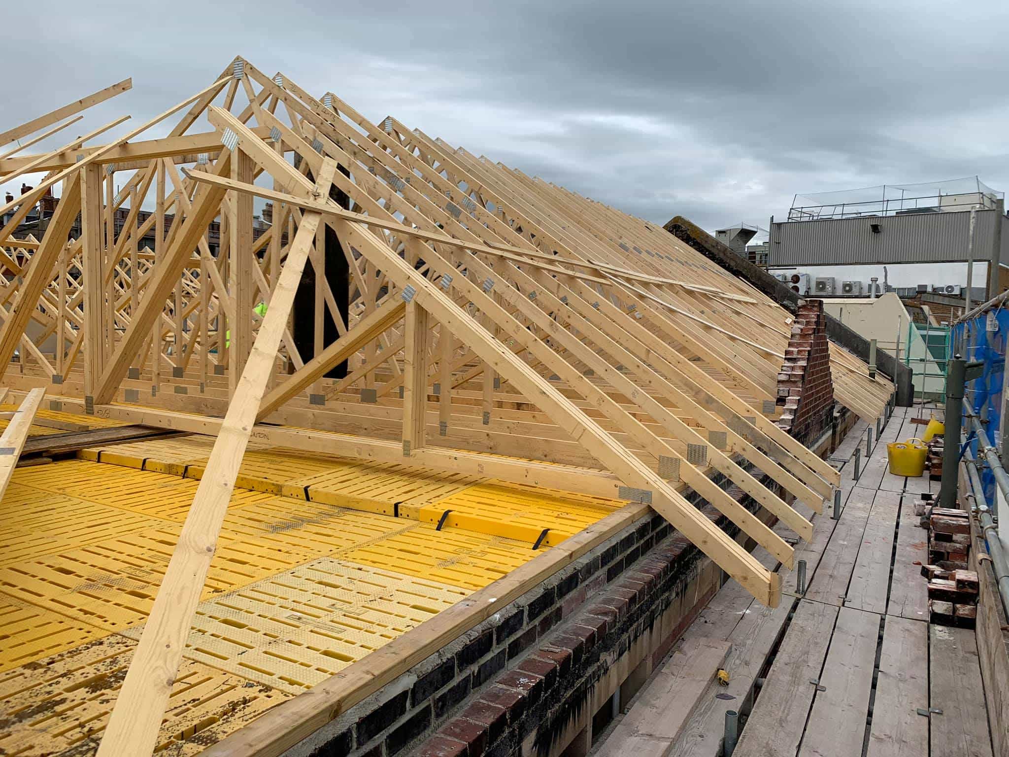 Wooden roof trusses and beams being installed on a building under construction, with scaffolding and tools visible on the right side under a cloudy sky.