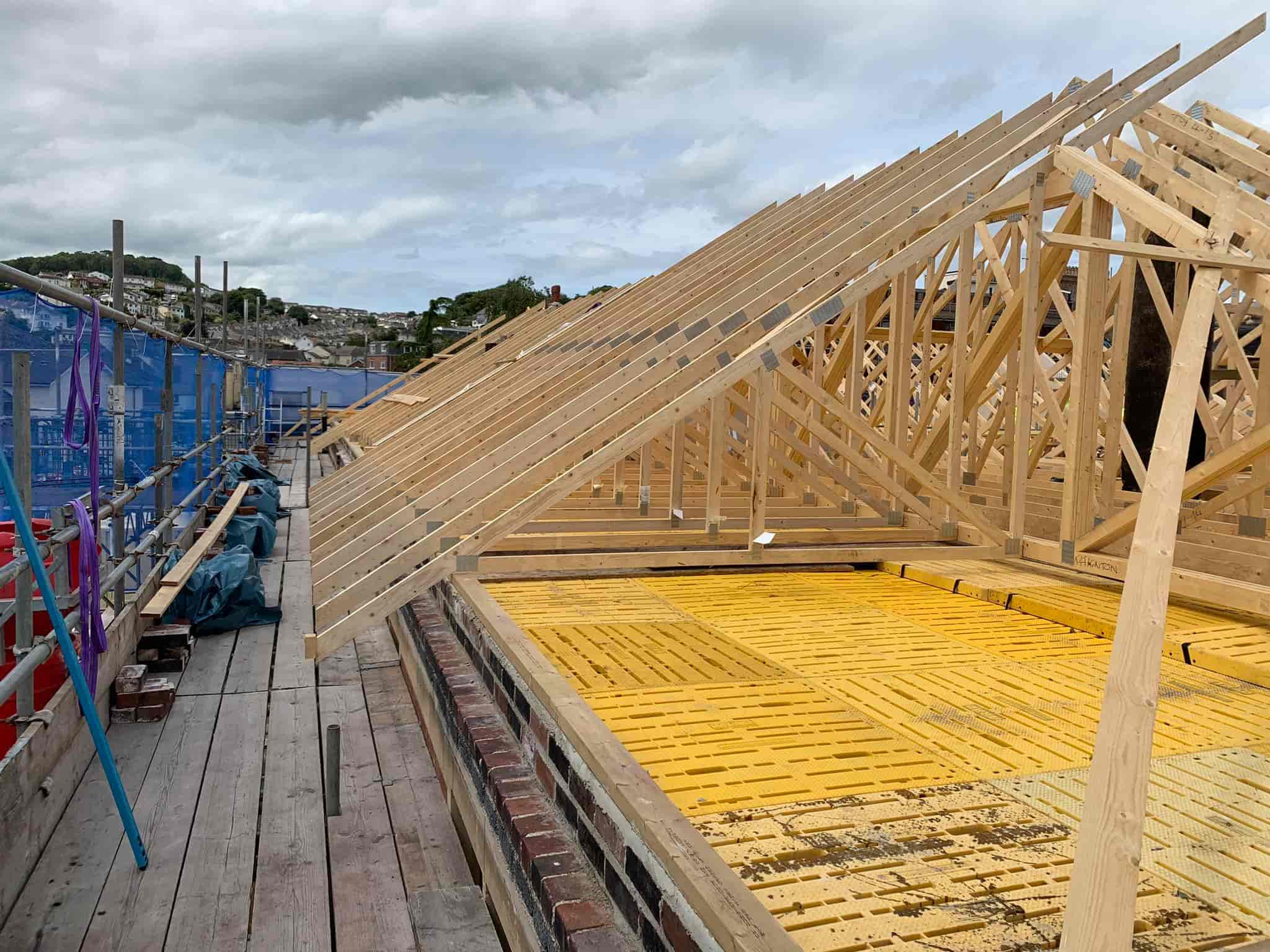 Partially constructed building with exposed wooden roof trusses and yellow floor panels, surrounded by scaffolding and safety netting, under a cloudy sky.