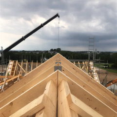 A crane lifts materials above a roof under construction, with wooden trusses in the foreground and scaffolding around the building. Overcast sky and green landscape are visible in the background.