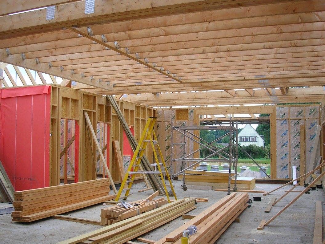 Wooden framing inside a house under construction, with exposed beams, stacked lumber, a yellow ladder, and scaffolding. Pink insulation and exterior sheathing are visible, with windows showing a house and greenery outside.