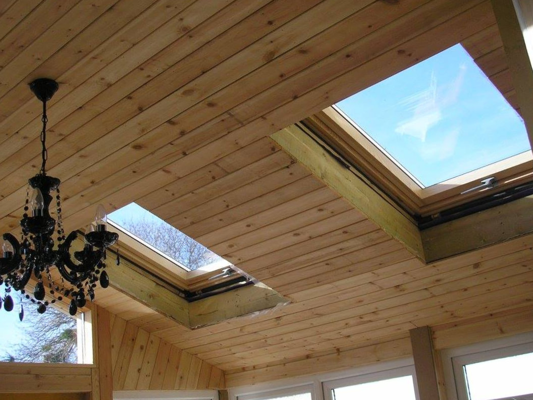 A wooden paneled ceiling with two large rectangular skylights allows natural light in. A black chandelier with hanging crystals is also visible in the left part of the image. Blue sky can be seen through the skylights.
