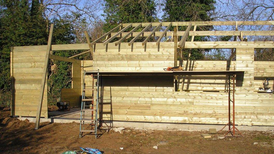 A wooden shed or small building under construction, with partial walls and roof framing in place. Scaffolding, tools, and materials are visible in front, and trees are in the background.