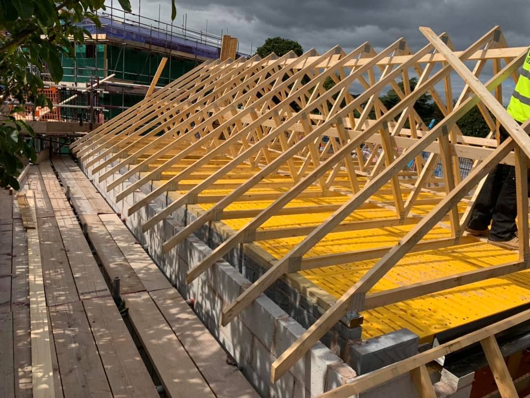Wooden roof trusses are being installed on top of a building under construction, with yellow boards covering the roof and scaffolding surrounding the site. A person in a hi-vis vest stands on the structure.