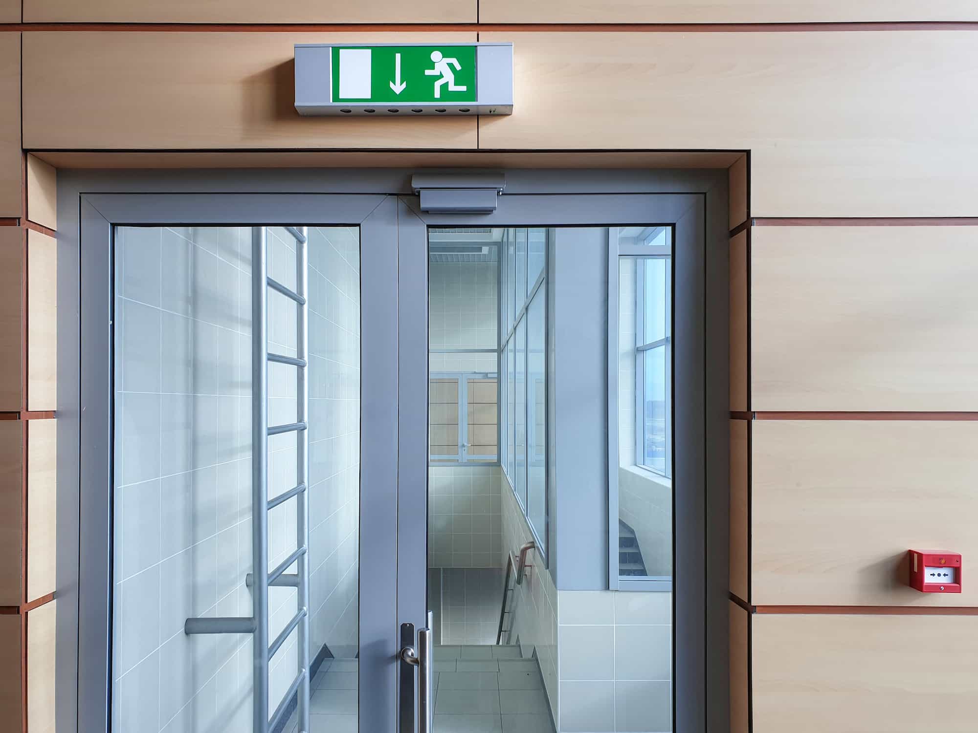 Glass double doors in a hallway with a green emergency exit sign above them and a red fire alarm on the wall to the right. The hallway appears clean and modern.