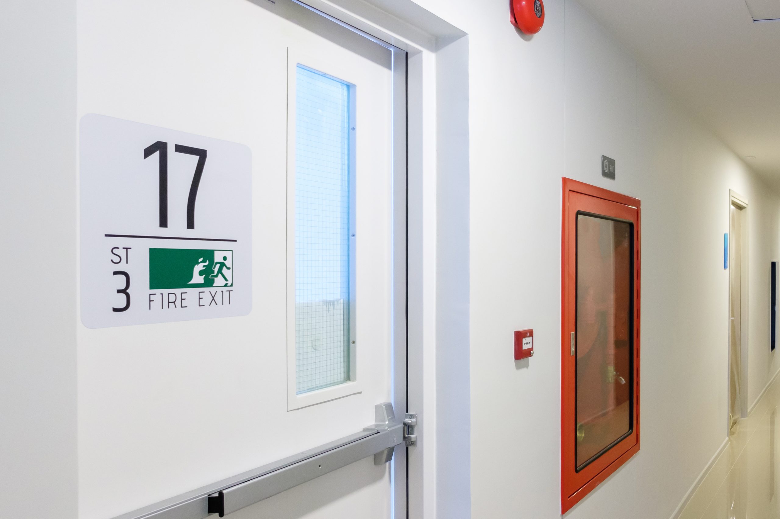 A white hallway with a door labeled "17 ST 3 Fire Exit" featuring an emergency exit sign. A red fire hose cabinet is mounted on the wall nearby, and the area is clean and well-lit.