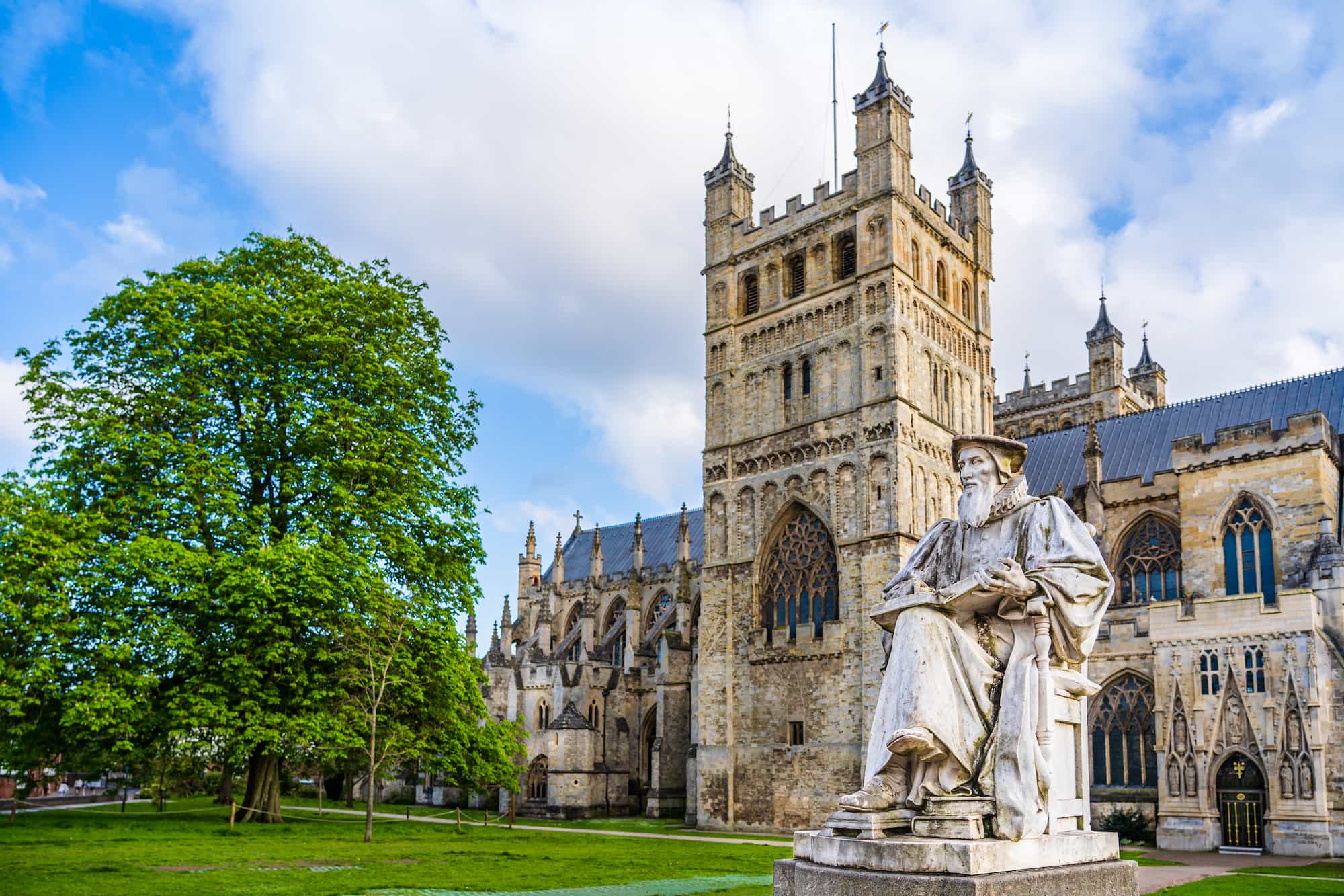A large stone statue of a seated figure stands in front of a historic Gothic cathedral with tall towers and arched windows, surrounded by green grass and a leafy tree under a partly cloudy sky.
