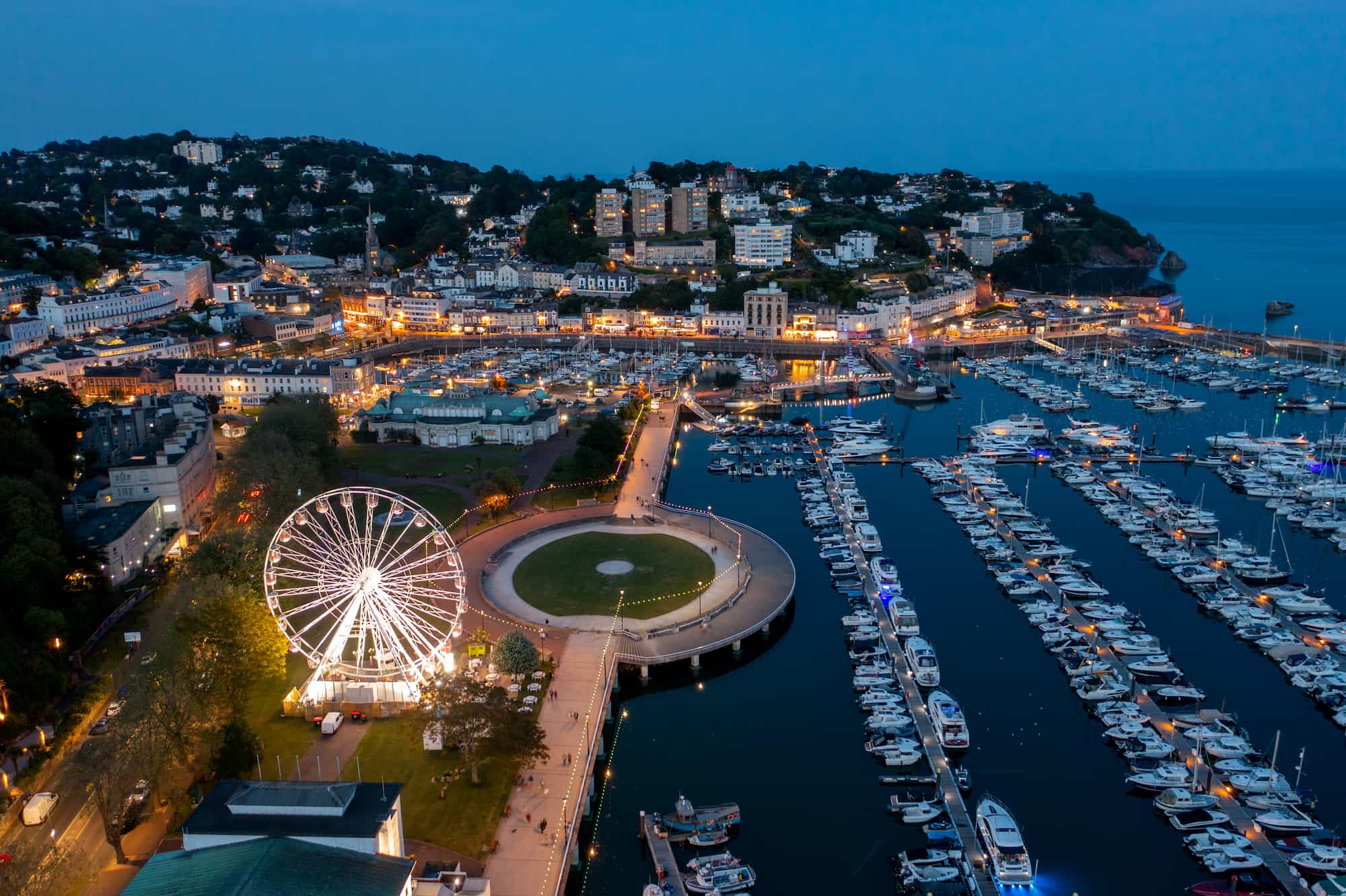 Aerial view of a marina at dusk with rows of docked boats, a large lit Ferris wheel, circular park, and surrounding town buildings illuminated by streetlights near the coast.