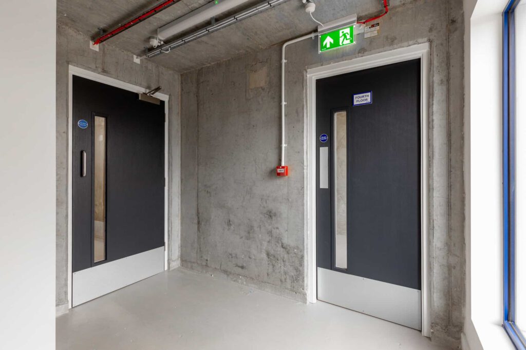 A concrete-walled corridor with two black fire doors, each with a vertical window. A green emergency exit sign with a running figure and arrow points above the right door, and a red fire alarm is mounted on the wall between the doors.