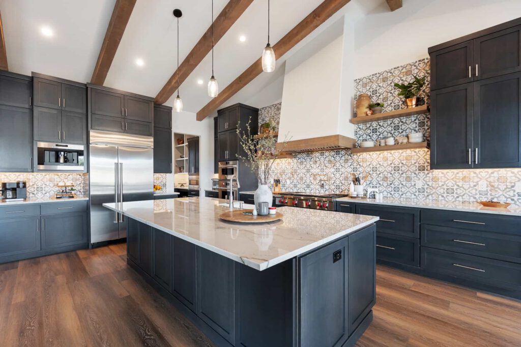 Modern kitchen with dark cabinets, white marble countertops, patterned tile backsplash, stainless steel appliances, and wooden ceiling beams. A large island with decor sits in the center on wooden flooring.