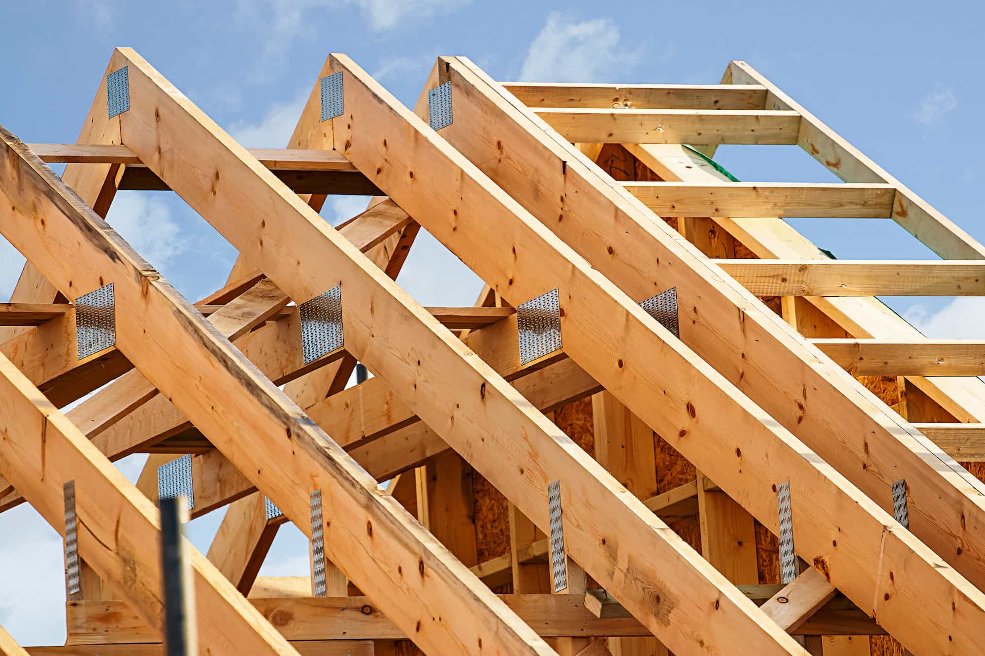 Close-up view of wooden roof trusses under construction, arranged diagonally against a blue sky with some clouds. The beams are exposed, showing metal connectors and unfinished wood.