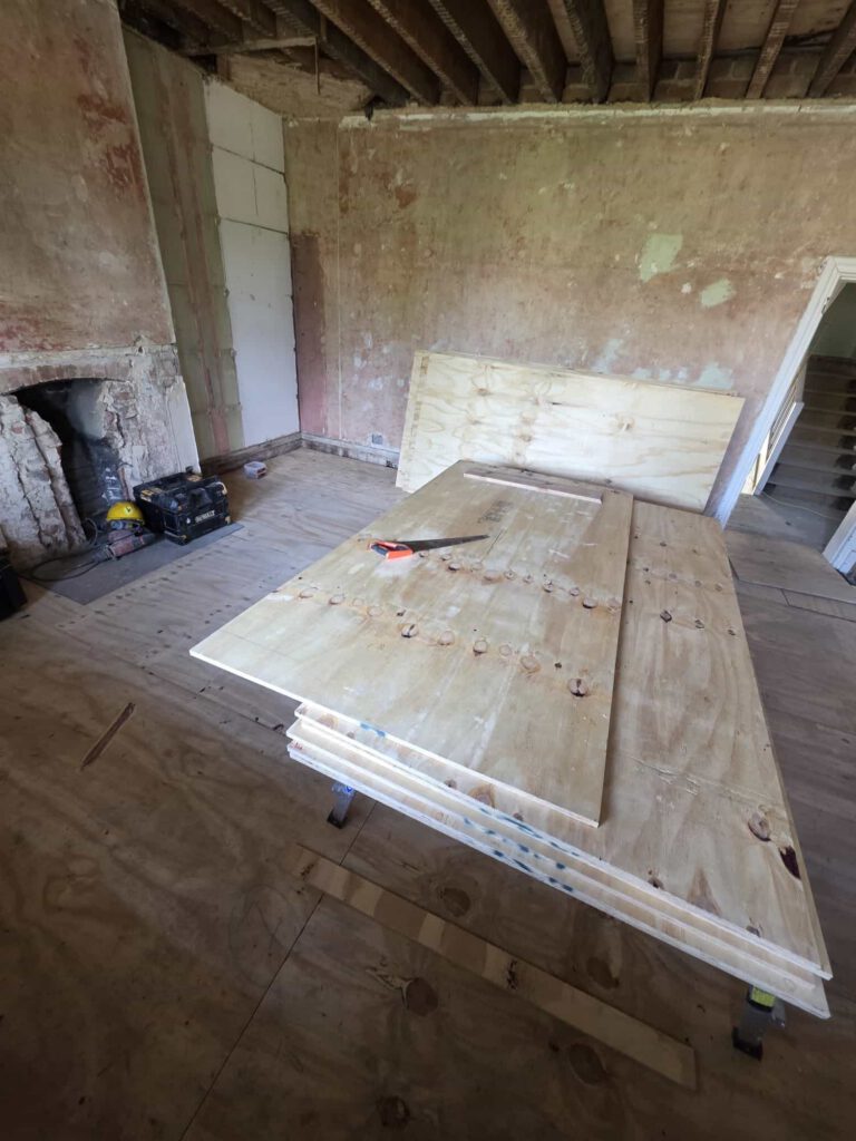 A room under renovation with exposed ceiling beams, unfinished walls, a fireplace, and several large plywood boards stacked on the floor. A handsaw rests on top of the plywood.