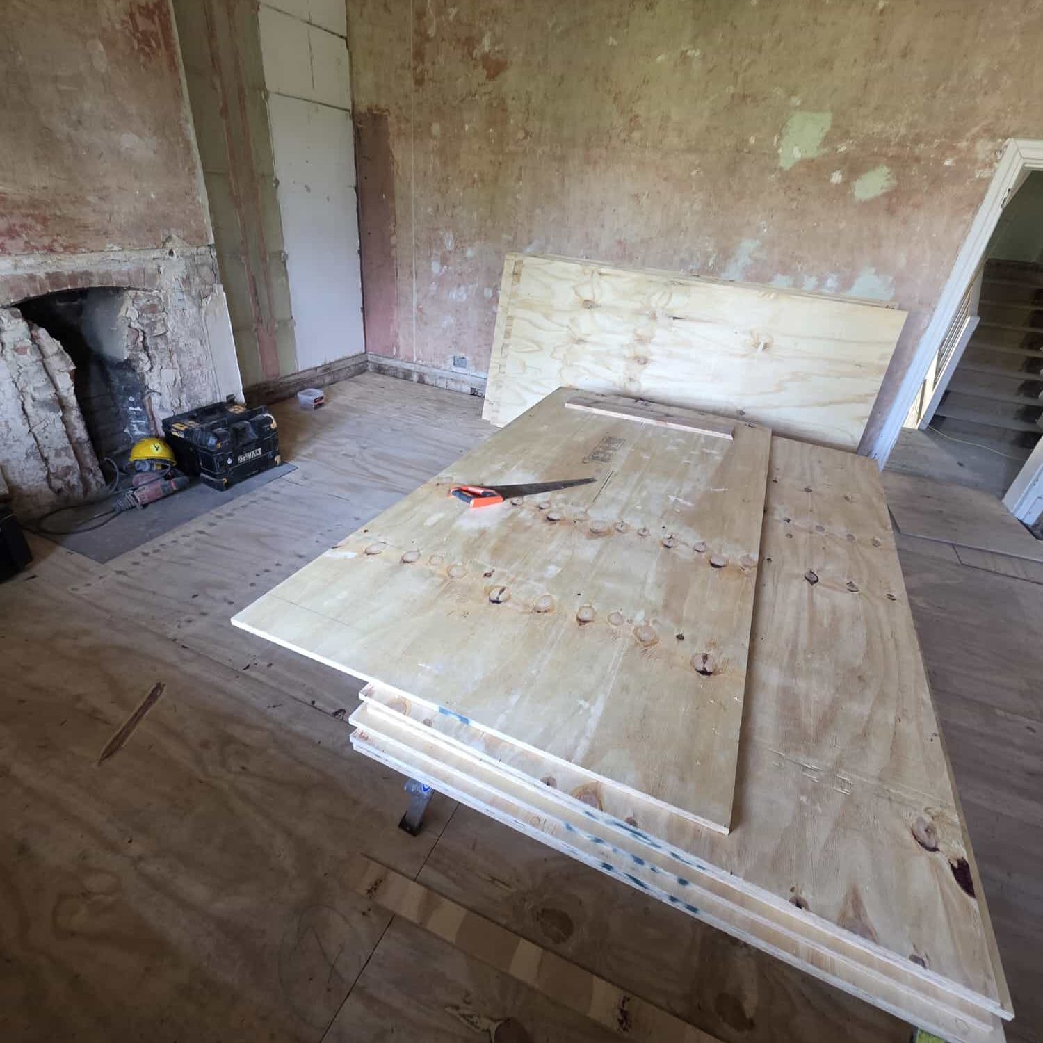 A room under renovation with unfinished plaster walls, plywood sheets stacked on the floor and a table, a saw on top, and construction tools near a bricked-up fireplace. Stairs and a doorway are visible in the background.
