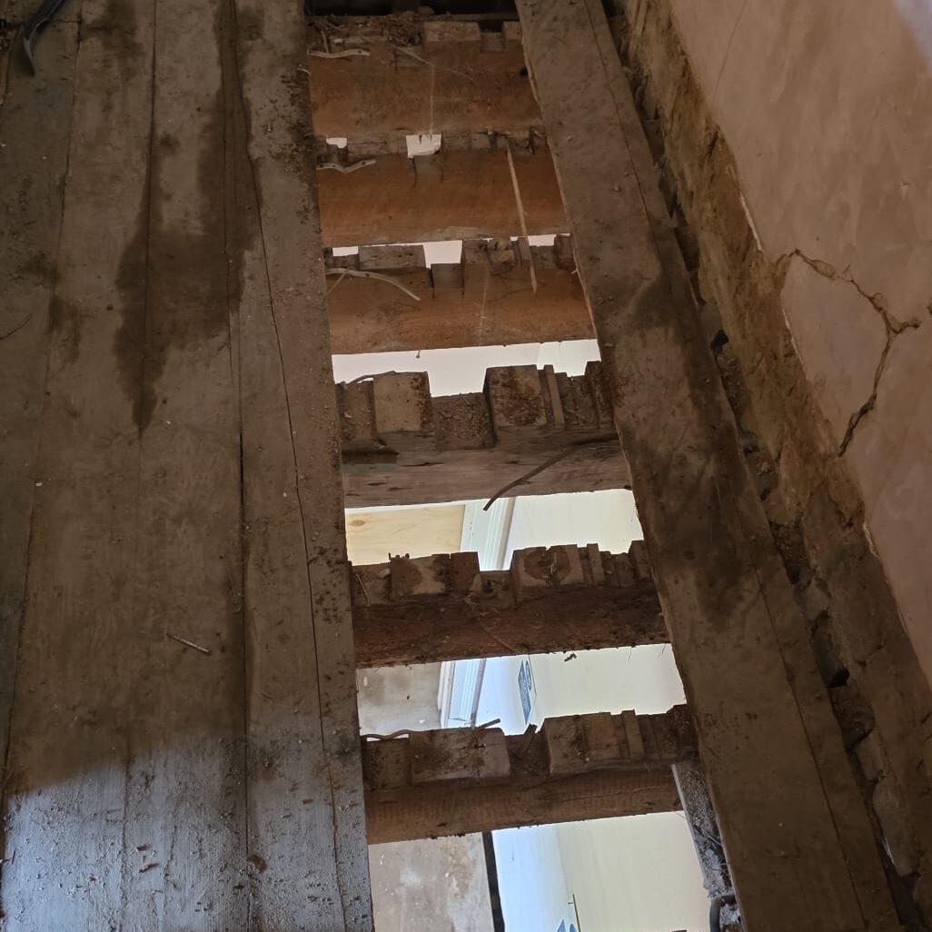 View from above of a partially demolished wooden floor, with exposed beams and gaps showing the room below. The wood is aged and damaged, and some plaster and brickwork are visible on the surrounding wall.