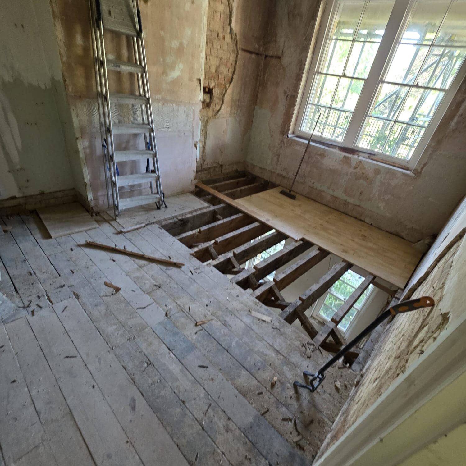 A room under renovation with floorboards partially removed, exposing wooden beams. A ladder leans against the wall, and a wooden plank covers part of the opening near a window. Tools are scattered on the floor.