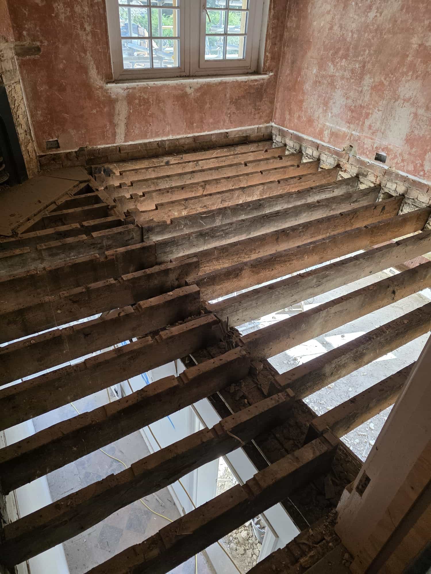 A room under renovation with exposed wooden floor joists and no floorboards, revealing the space below. The walls are unfinished, and sunlight enters through a window with white trim.