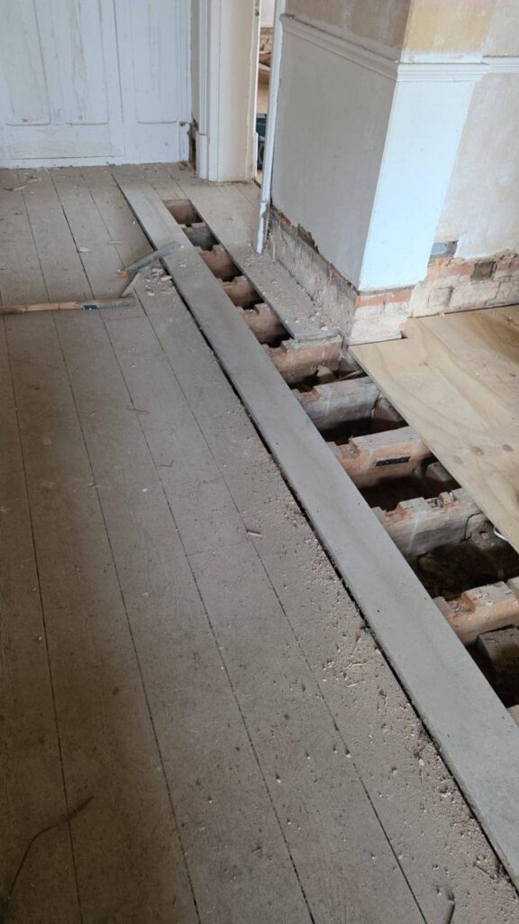 A wooden floor with several floorboards removed, exposing the joists and subfloor beneath. Some tools and debris are scattered around, suggesting ongoing renovation work.