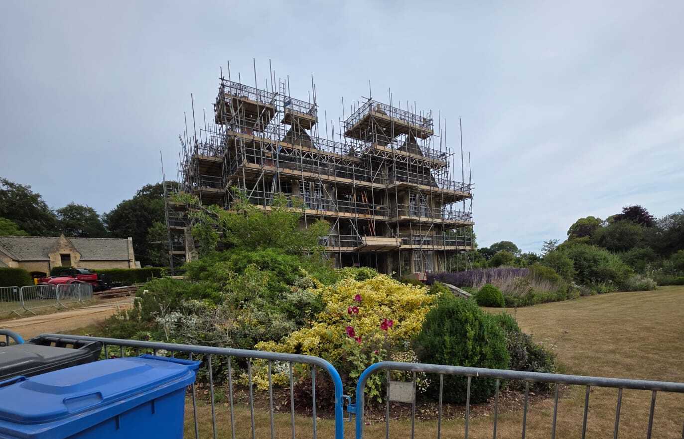 A large building covered in scaffolding stands behind metal barriers and lush gardens, with a blue recycling bin visible in the foreground under a cloudy sky.