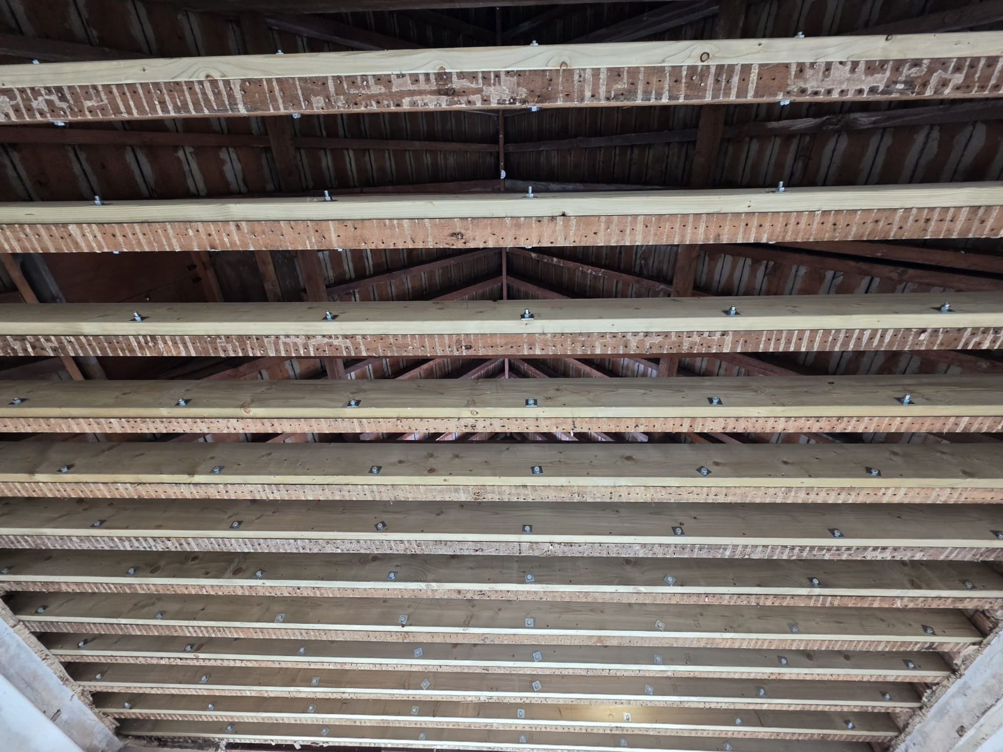 View of a wooden ceiling framework with evenly spaced horizontal wooden beams and visible metal bolts, showing the underside of a roof under construction.