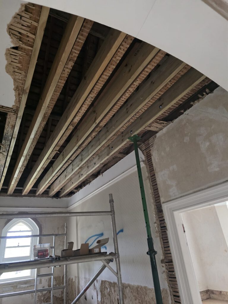A room under renovation with exposed wooden ceiling beams, scaffolding, a green support pole, and construction materials. The walls are partially stripped, and a window with an arch lets in daylight.