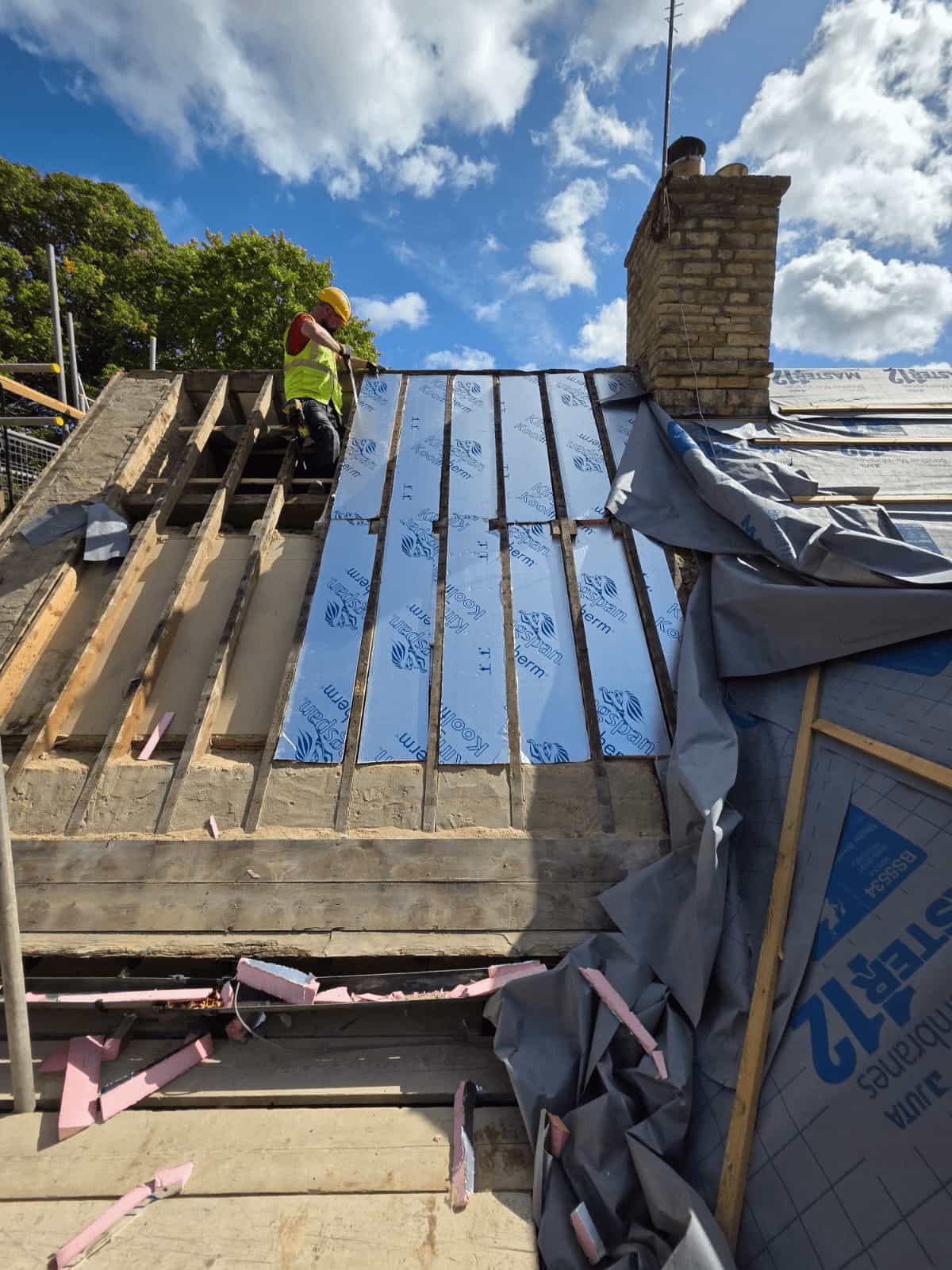 A construction worker in a yellow vest stands on a partially roofed building, working near a chimney under a partly cloudy blue sky. Some roof panels and insulation material are exposed.