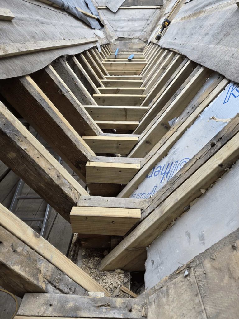View of a partially constructed roof structure with exposed wooden beams and supports, forming a V-shaped channel; surrounding materials and some insulation covers are visible on the sides.