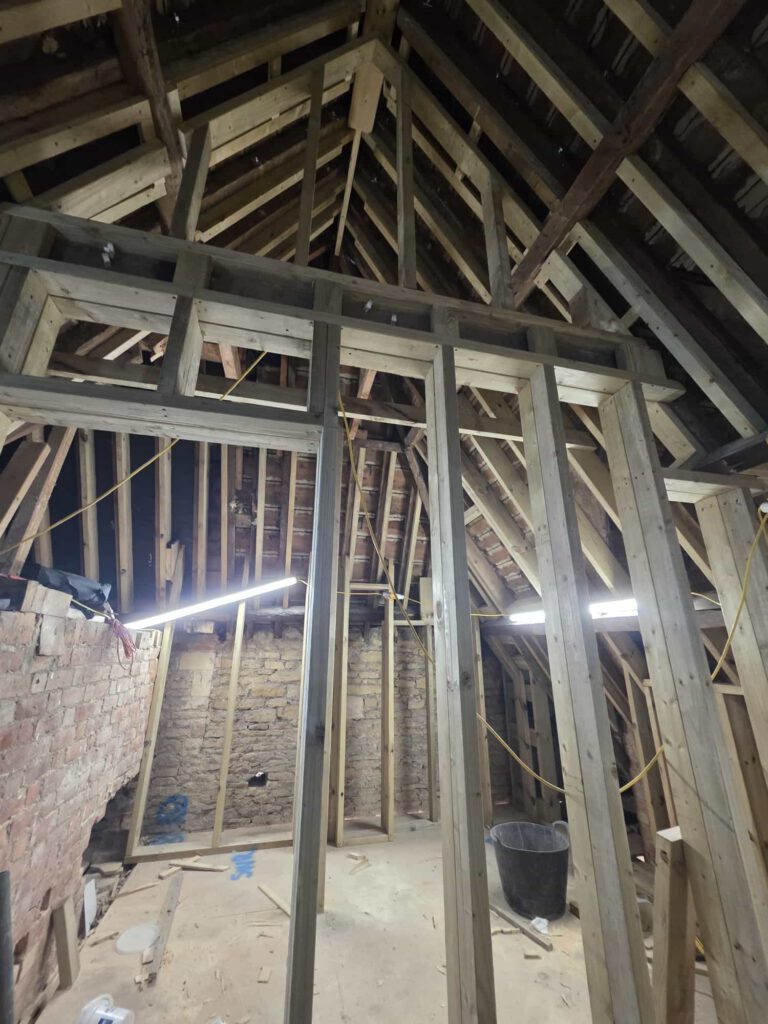 An unfinished attic space with exposed wooden beams, studs, and rafters. Construction materials and tools are scattered on the floor. Bright work lights hang from the ceiling, illuminating the area.