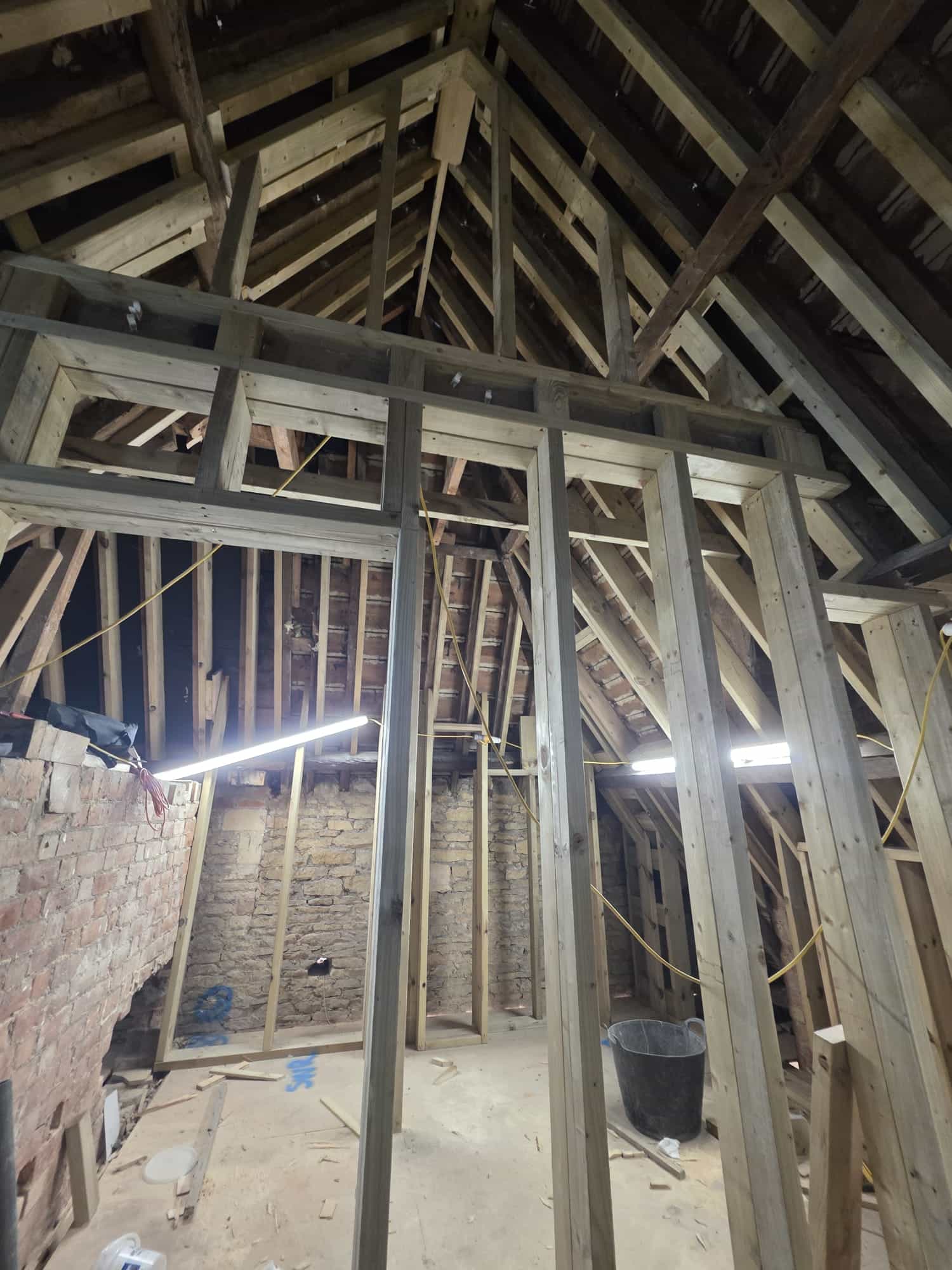 An unfinished attic space with exposed wooden beams, studs, and rafters. Construction materials and tools are scattered on the floor. Bright work lights hang from the ceiling, illuminating the area.