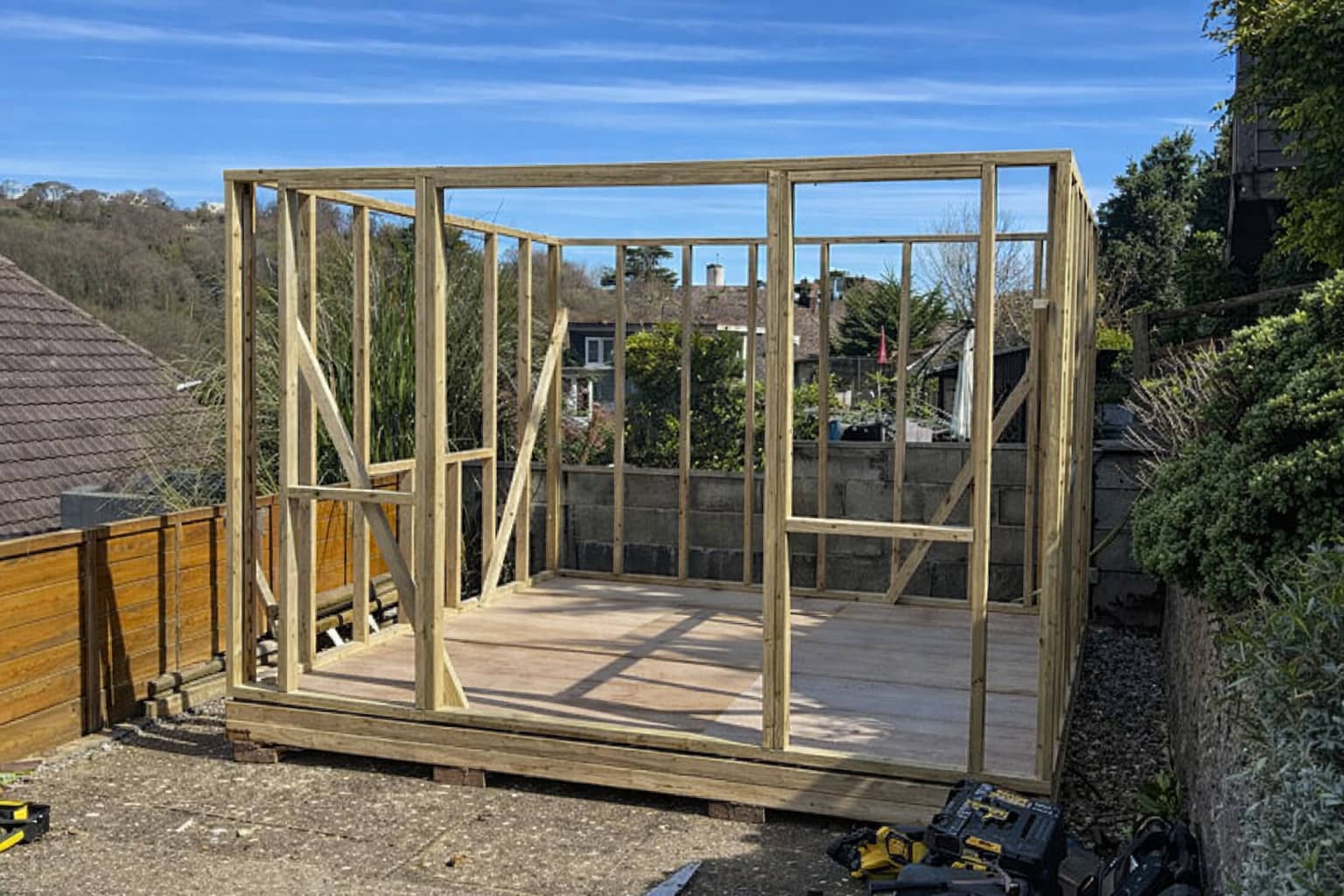 A wooden shed frame under construction stands on a concrete base outdoors, surrounded by garden greenery and nearby houses, under a blue sky with wispy clouds. Tools are visible on the ground.