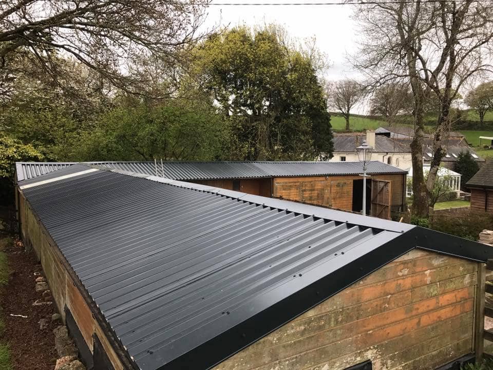 A view of buildings with corrugated metal roofs surrounded by trees and greenery, with other houses and open fields visible in the background. The scene appears to be in a rural area.
