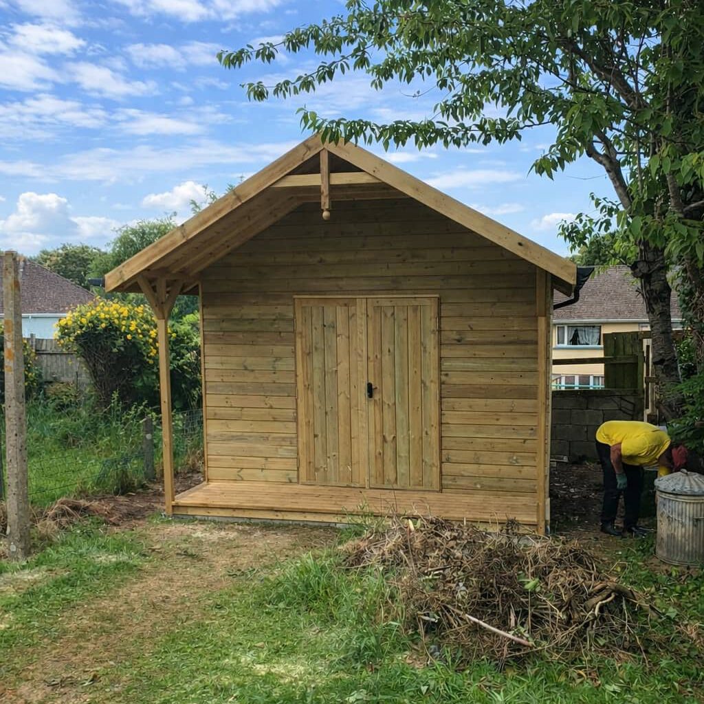 A wooden garden shed with a small porch stands on a grassy yard near trees and a house. A person in a yellow shirt is bending over next to a metal bin on the right side of the shed.