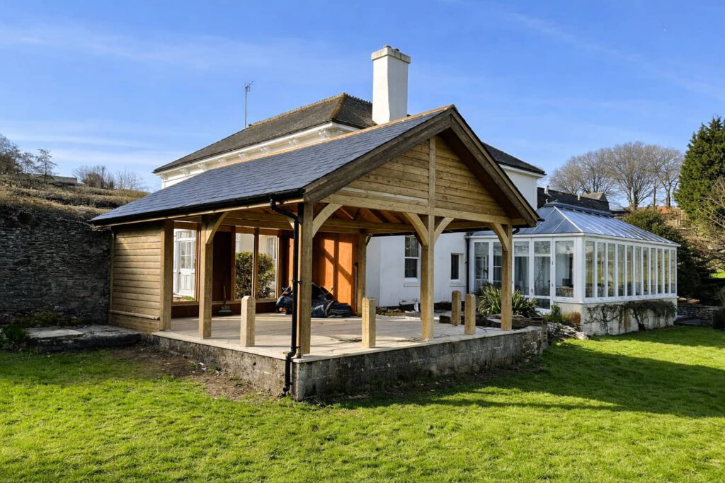 A wooden gazebo structure with a slate roof stands on a raised concrete base beside a white house with a glass conservatory, surrounded by green grass and trees under a clear blue sky.