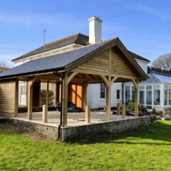 A wooden gazebo structure with a slate roof stands on a raised concrete base beside a white house with a glass conservatory, surrounded by green grass and trees under a clear blue sky.