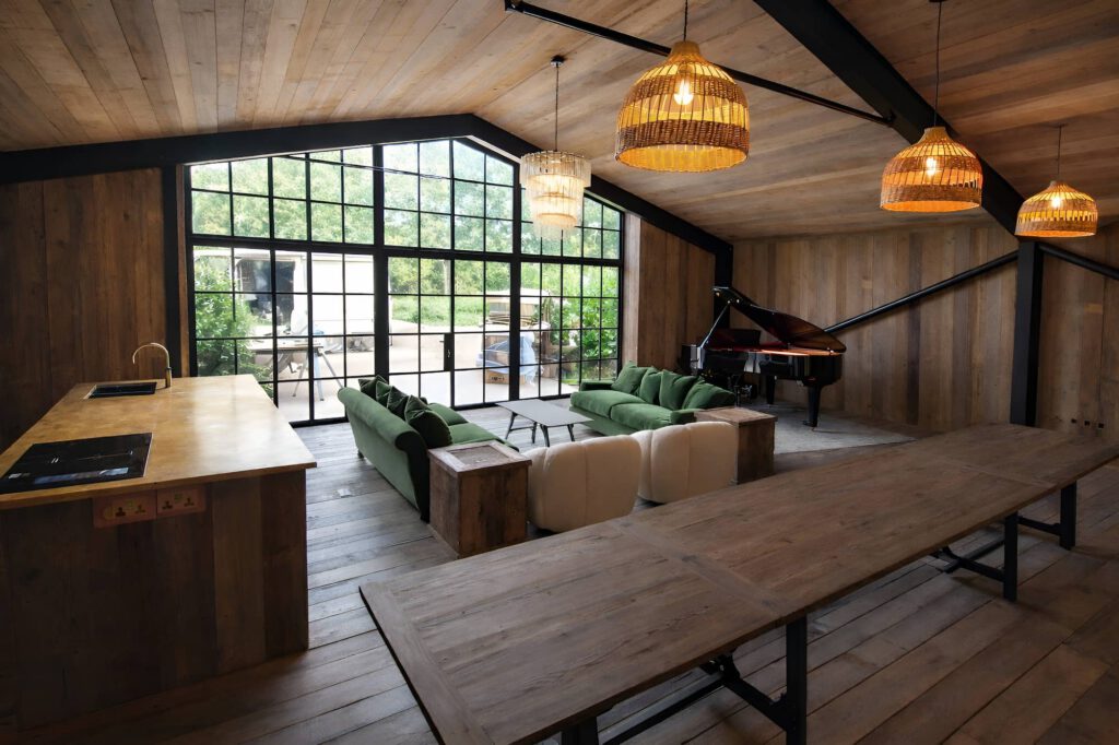 Spacious, modern living area with wood floors and ceilings, large windows, green sofas, a black grand piano, a long dining table, and pendant lights. Natural light fills the room, with greenery visible outside.