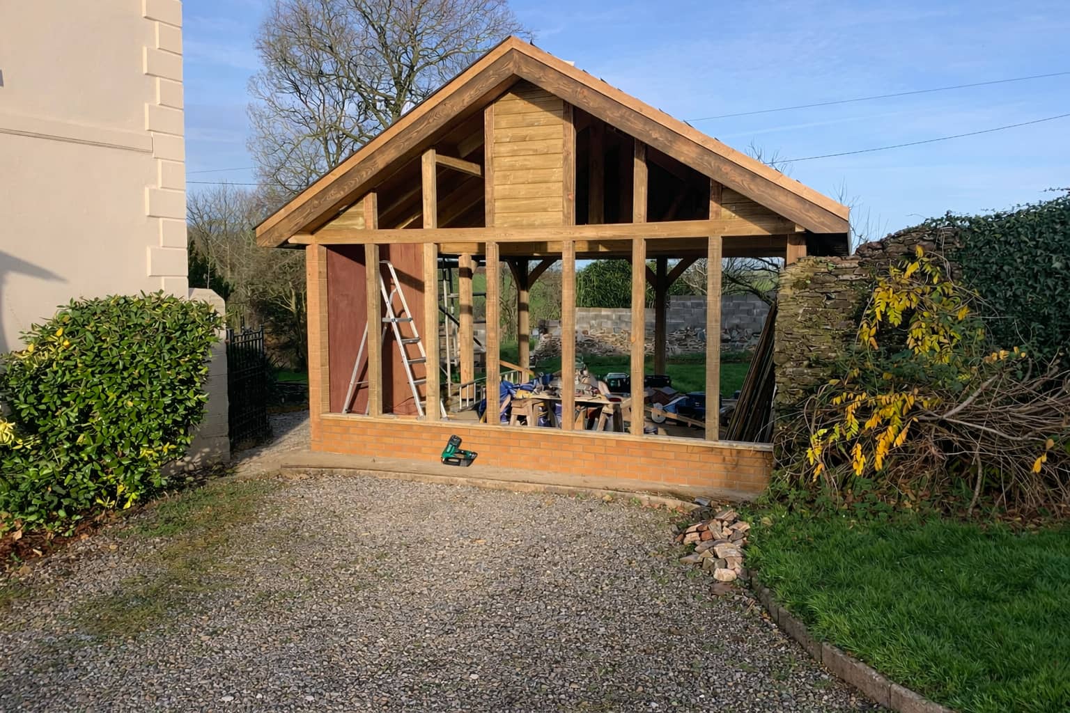 A wooden shed under construction with exposed framework, a pitched roof, and no windows or doors installed yet. A ladder and construction tools are inside, and there are bushes and grass nearby.