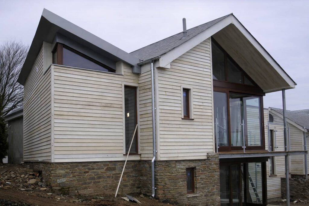A modern two-story house with light wooden siding, large glass windows, and a stone foundation, situated on uneven ground with construction tools nearby.